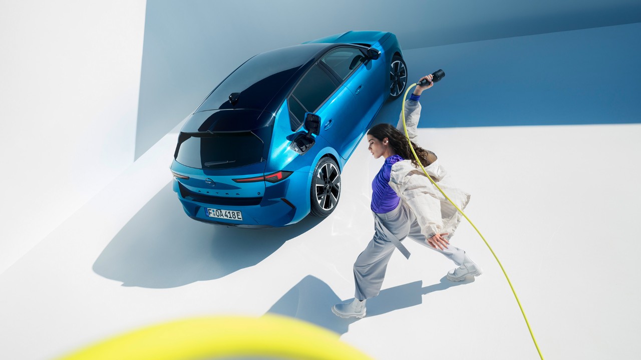 Vista de cima de um Opel Astra Electric azul com tejadilho preto, com uma mulher a rodar um cabo de carregamento amarelo