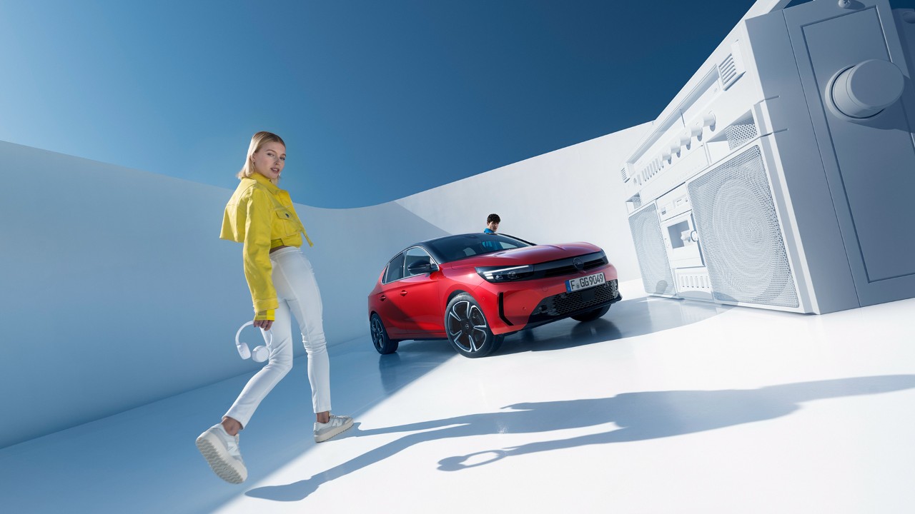 Mulher junto a novo Opel Corsa de cor vermelha com tejadilho preto e um homem próximo da porta do condutor